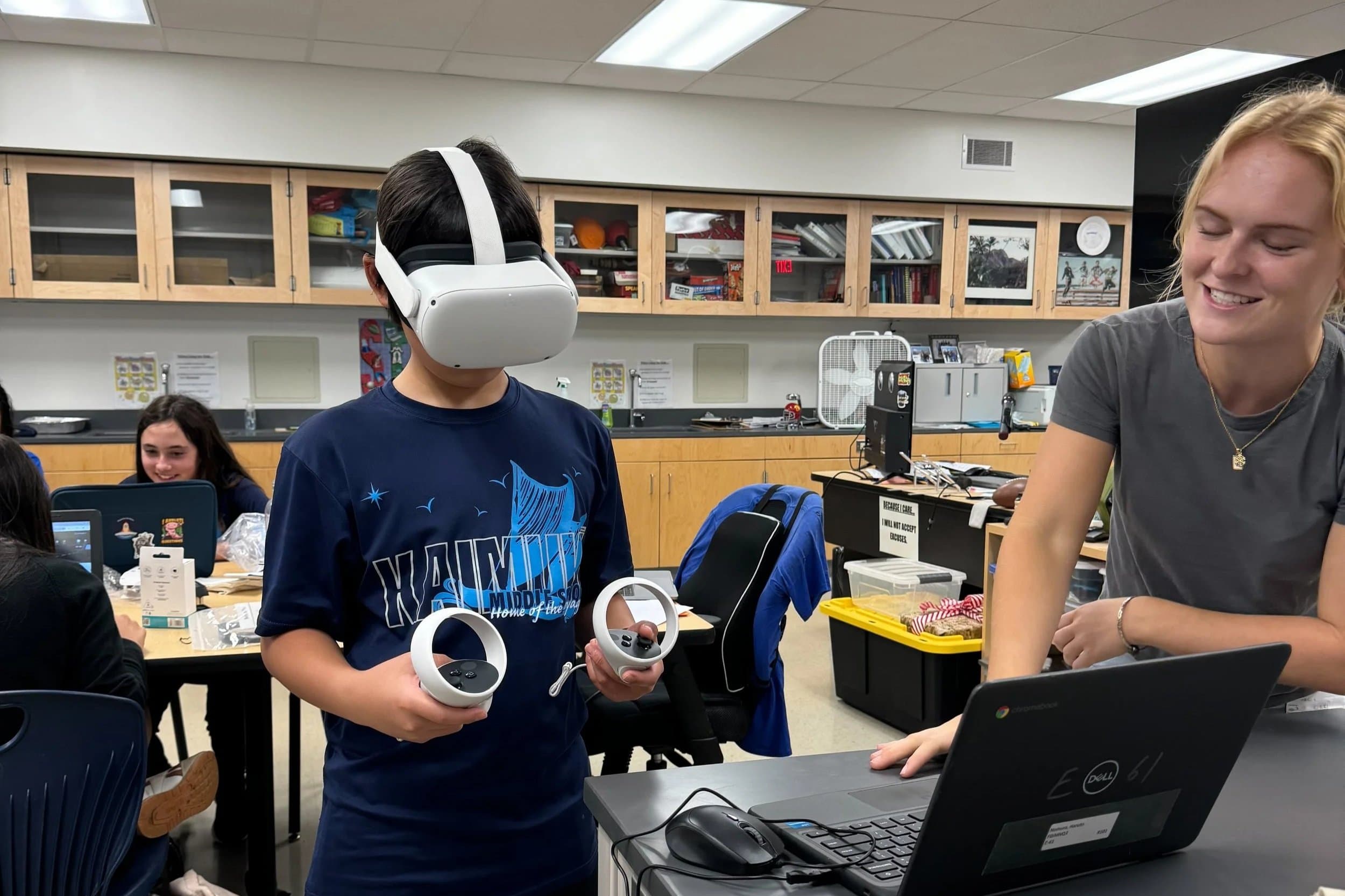 Student wearing a virtual reality headset and holding controllers in a classroom setting, with a teacher or facilitator smiling nearby, and other students working at tables in the background.