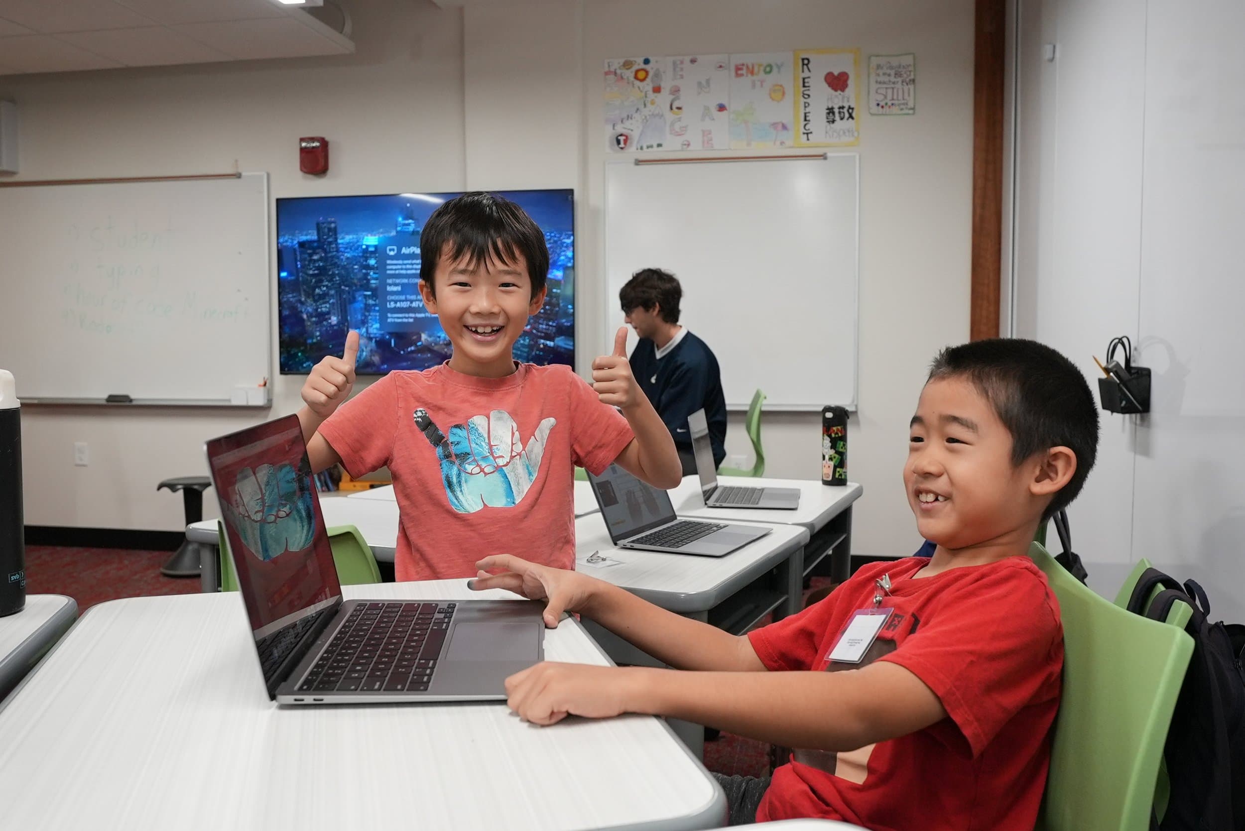 Two young boys smiling and giving thumbs-up in a classroom with laptops on the desks. A third person is working in the background, with educational posters on the wall.