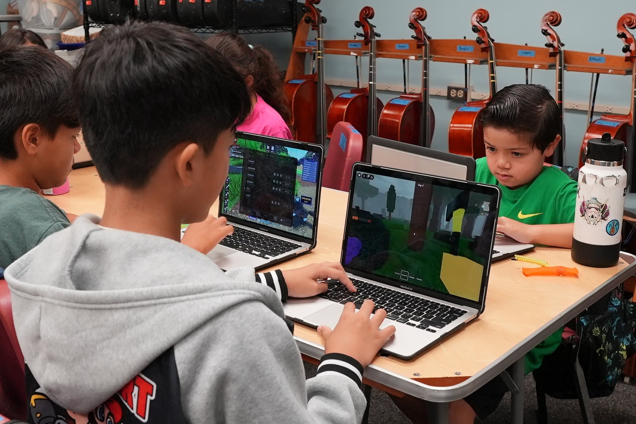 Four children playing video games on laptops at a classroom table with string instruments hanging on the wall behind them.