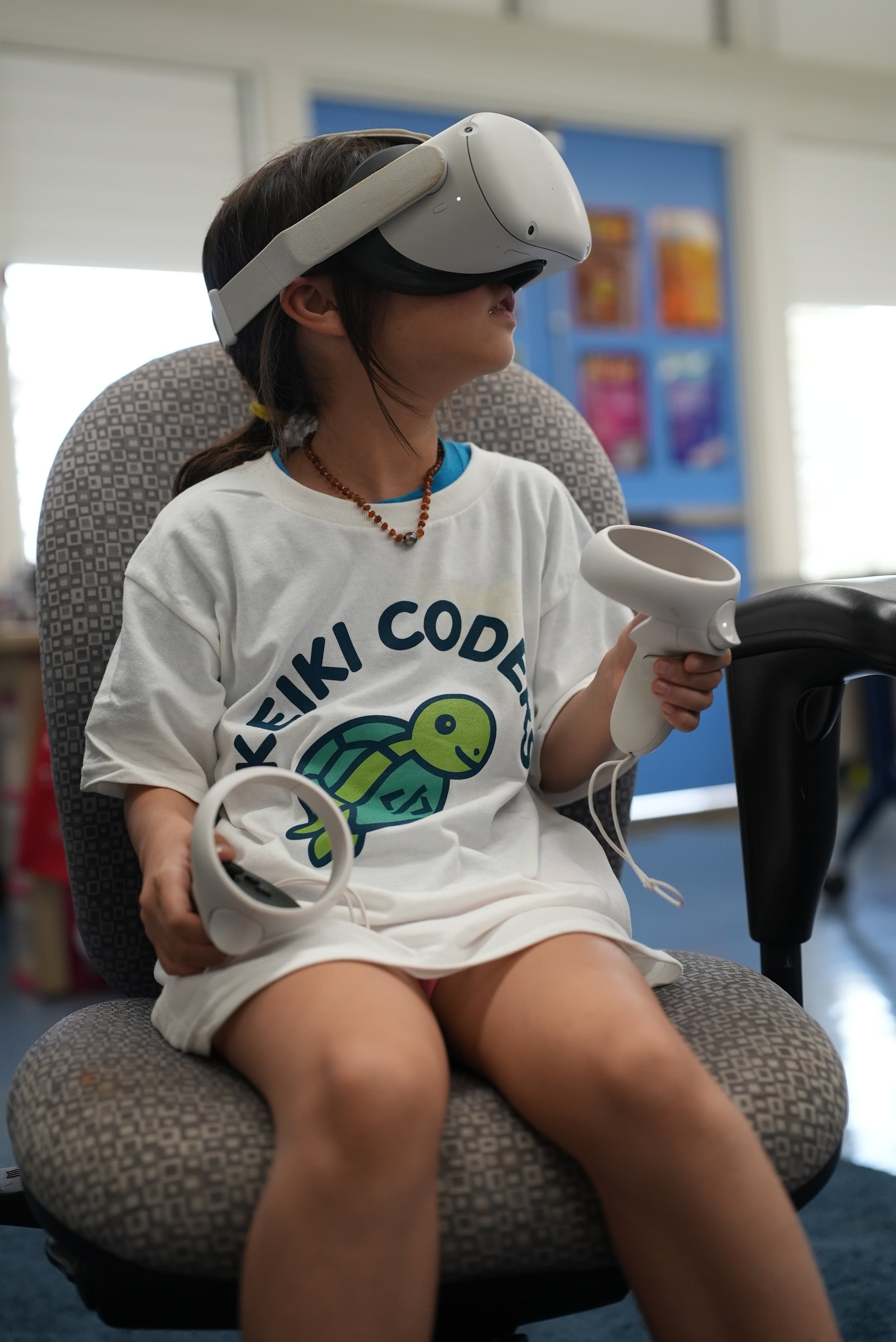 A young girl with dark hair and a beaded necklace sitting on a patterned chair, wearing a white T-shirt with a turtle graphic, using a virtual reality headset and holding a VR controller in her hand.