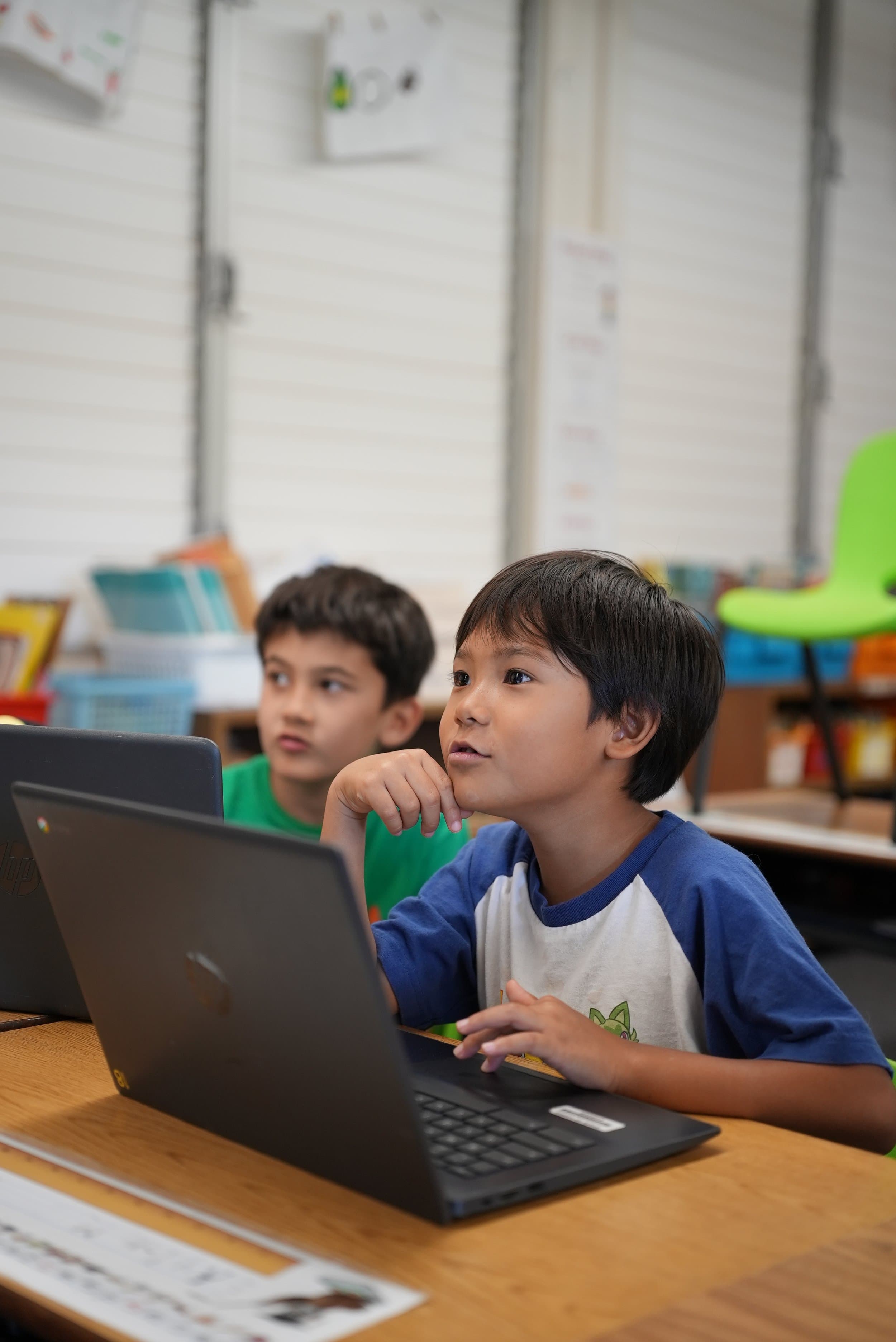 Two children working on Chromebook laptops in a classroom, the one in front resting their chin on a hand.
