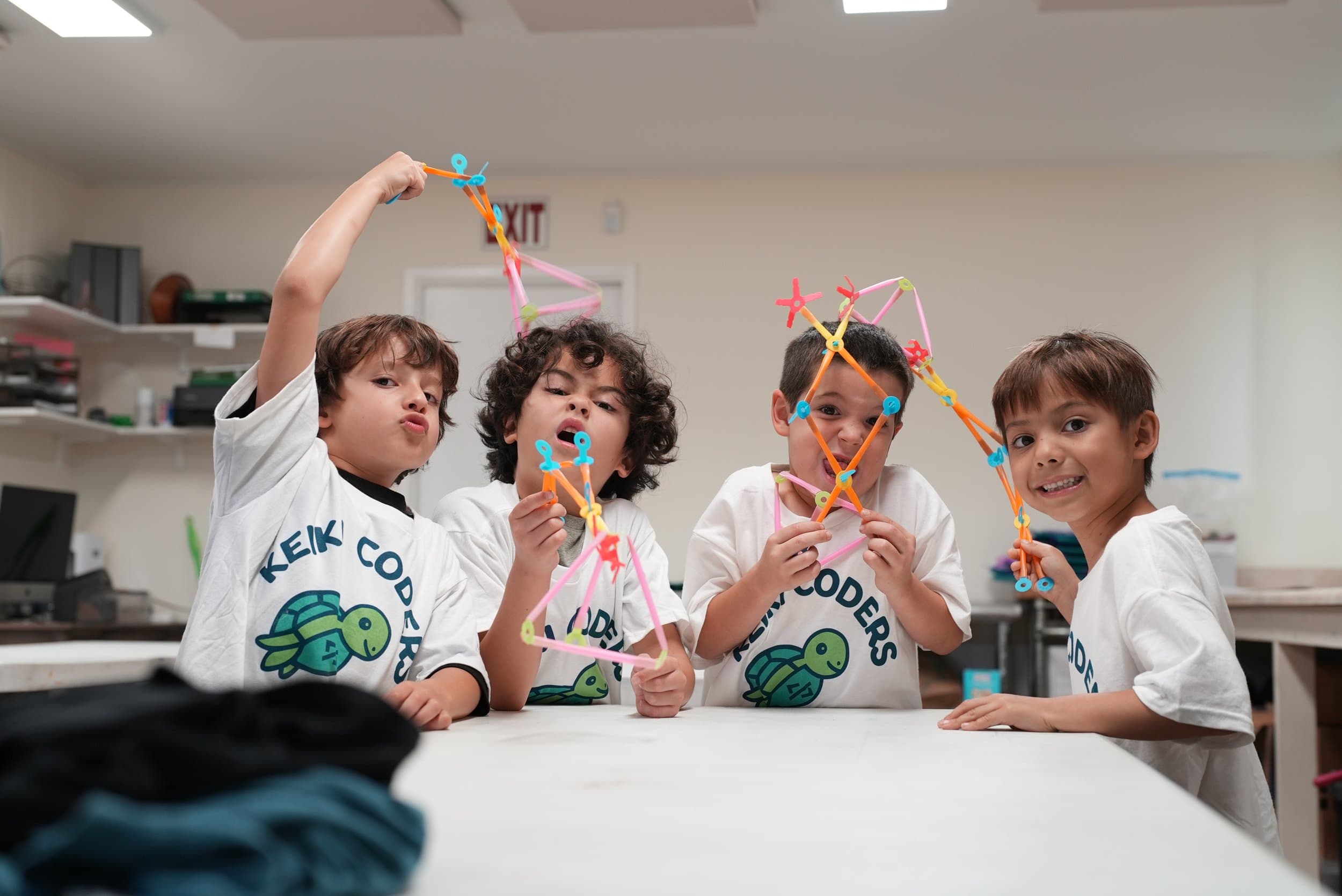 Four children wearing white T-shirts with a turtle graphic, holding colorful geometric shapes and posing for the camera in a classroom.