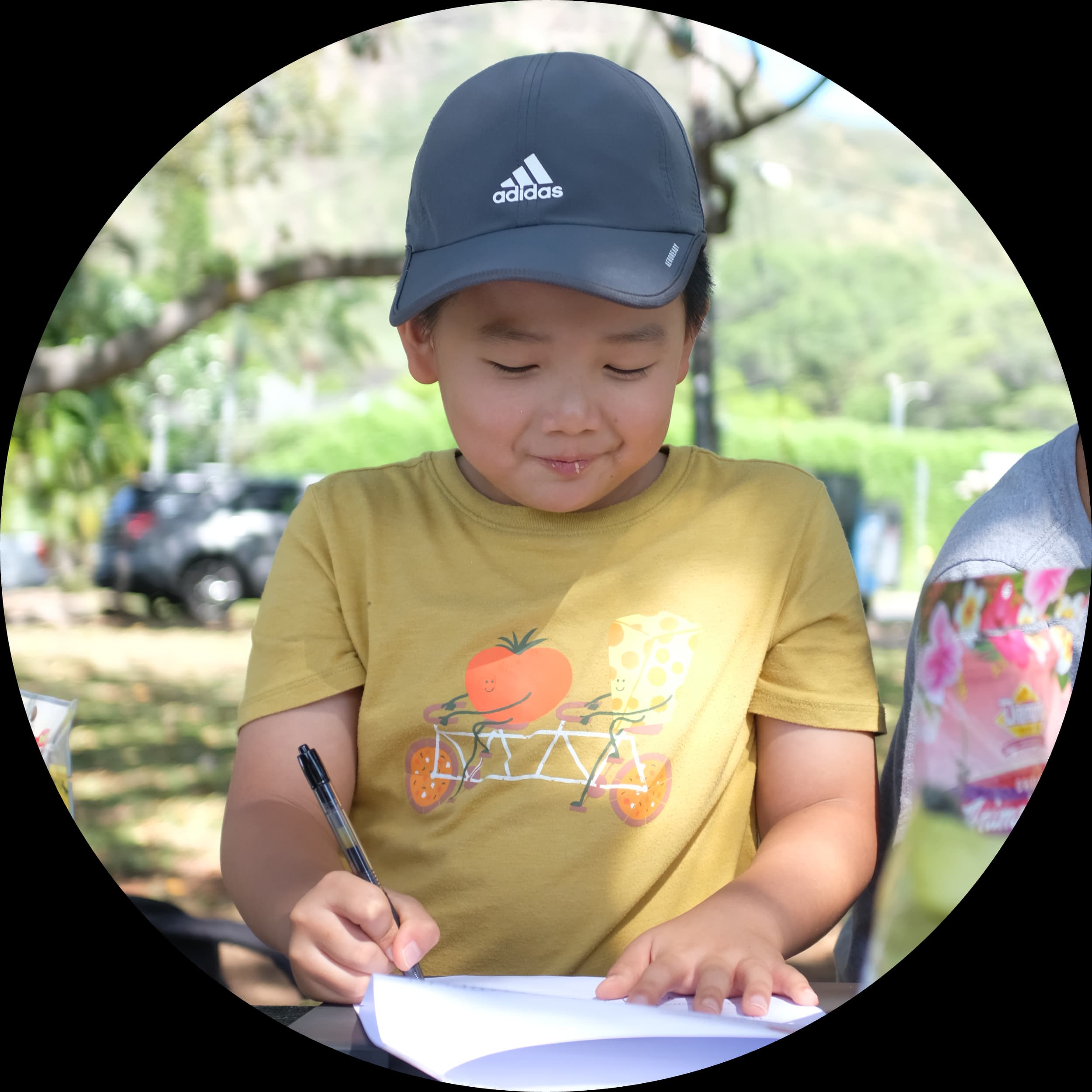 A young boy wearing a blue Adidas cap and a yellow T-shirt with a graphics of a tomato and cheese riding a bicycle, writing in a notebook outdoors during daytime.