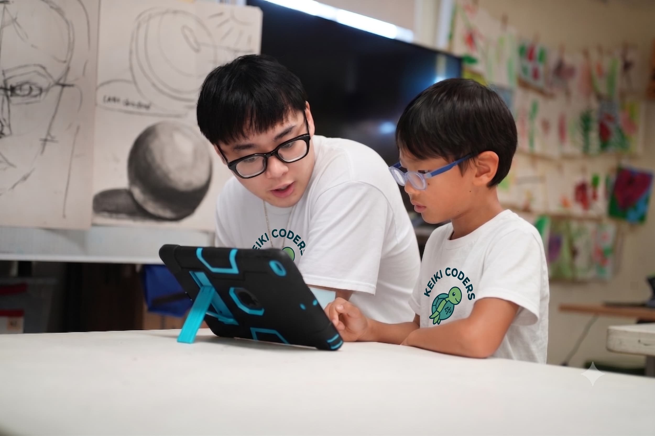 Two boys wearing glasses and white T-shirts with a turtle logo are sitting at a table, focused on a device or project.