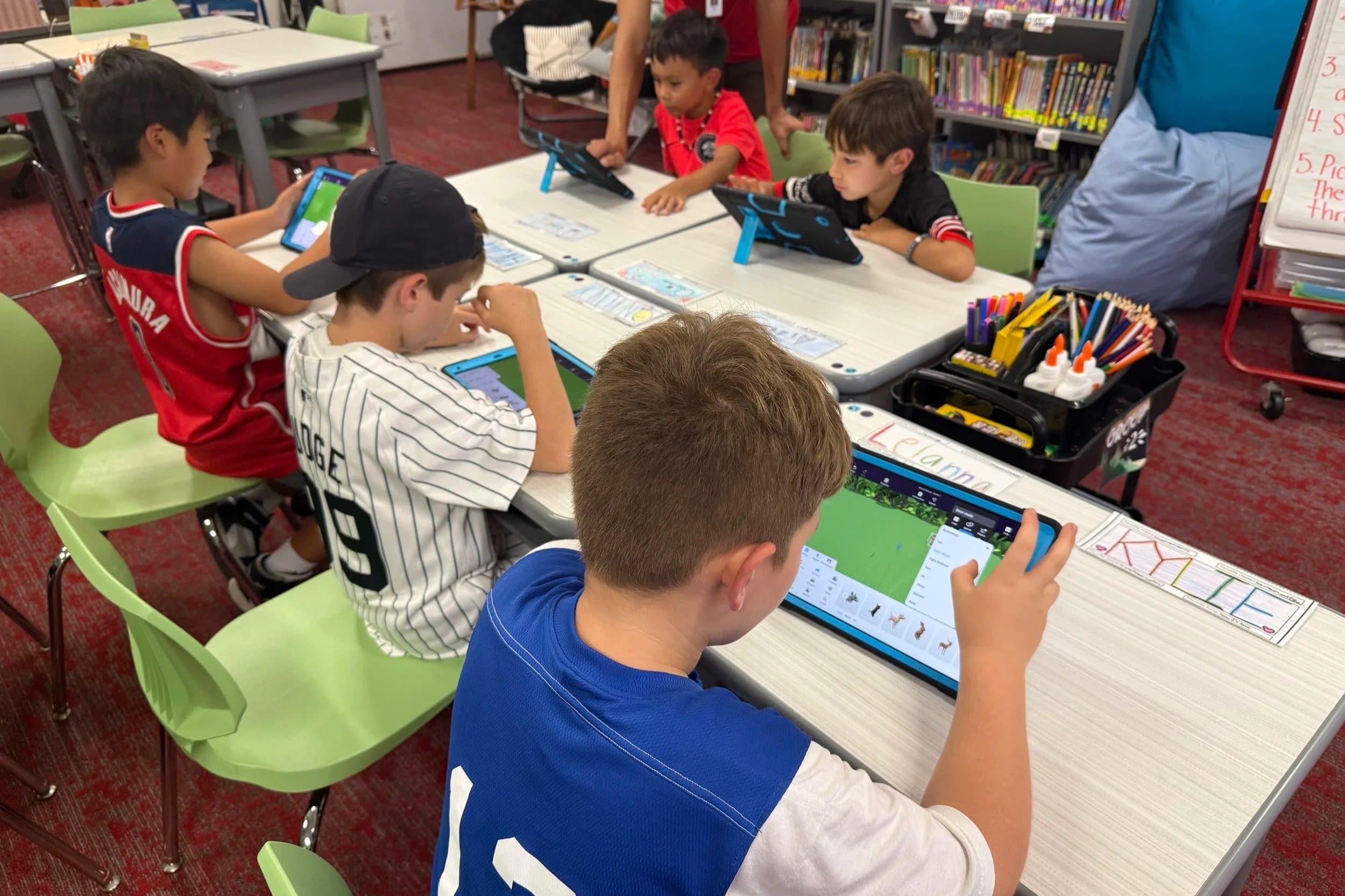Six children sitting at a table in a classroom, using tablets for educational activities. There is a library bookshelf in the background, and various school supplies on the table.