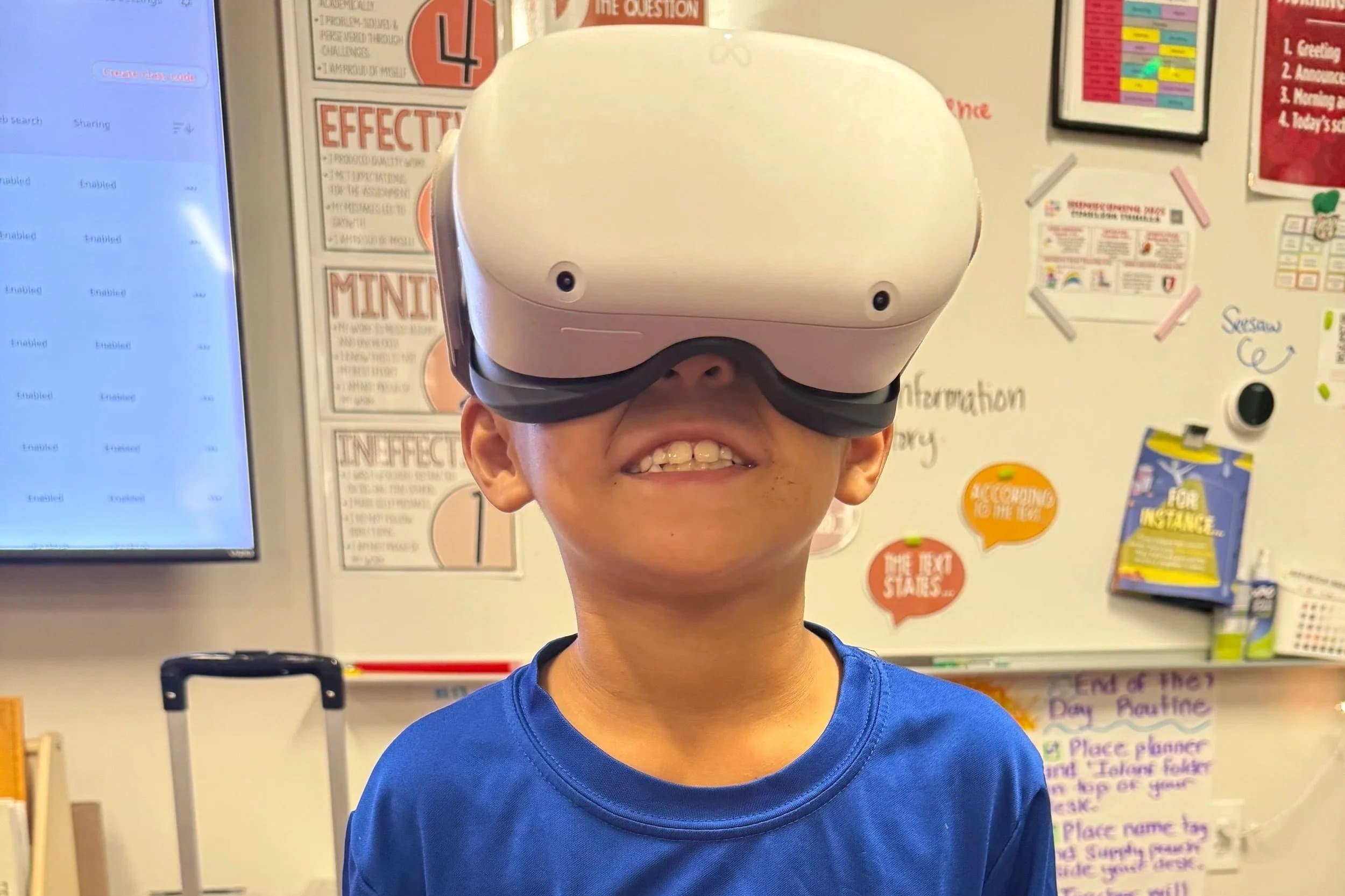 A young boy wearing a blue shirt and a virtual reality headset, smiling, in a classroom setting with educational posters and bulletin boards in the background.