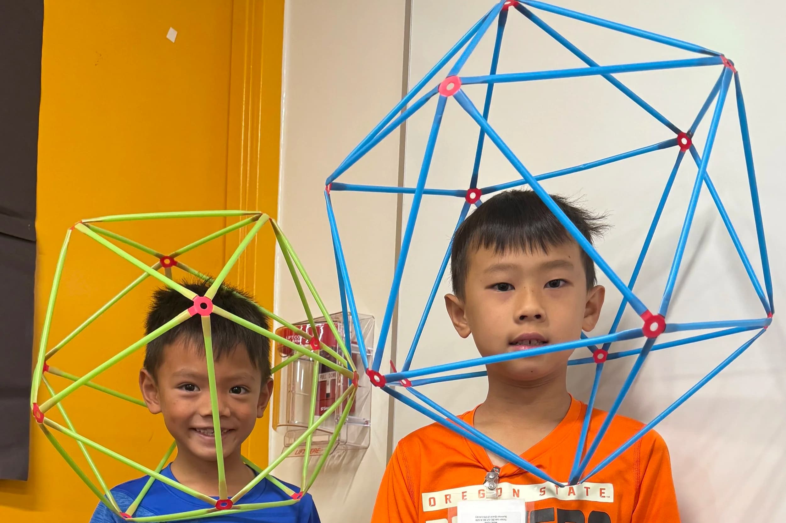 Two young boys standing with large geometric frame structures, one yellow and one blue. The boys are smiling and looking at the camera, inside a room with yellow and white walls.