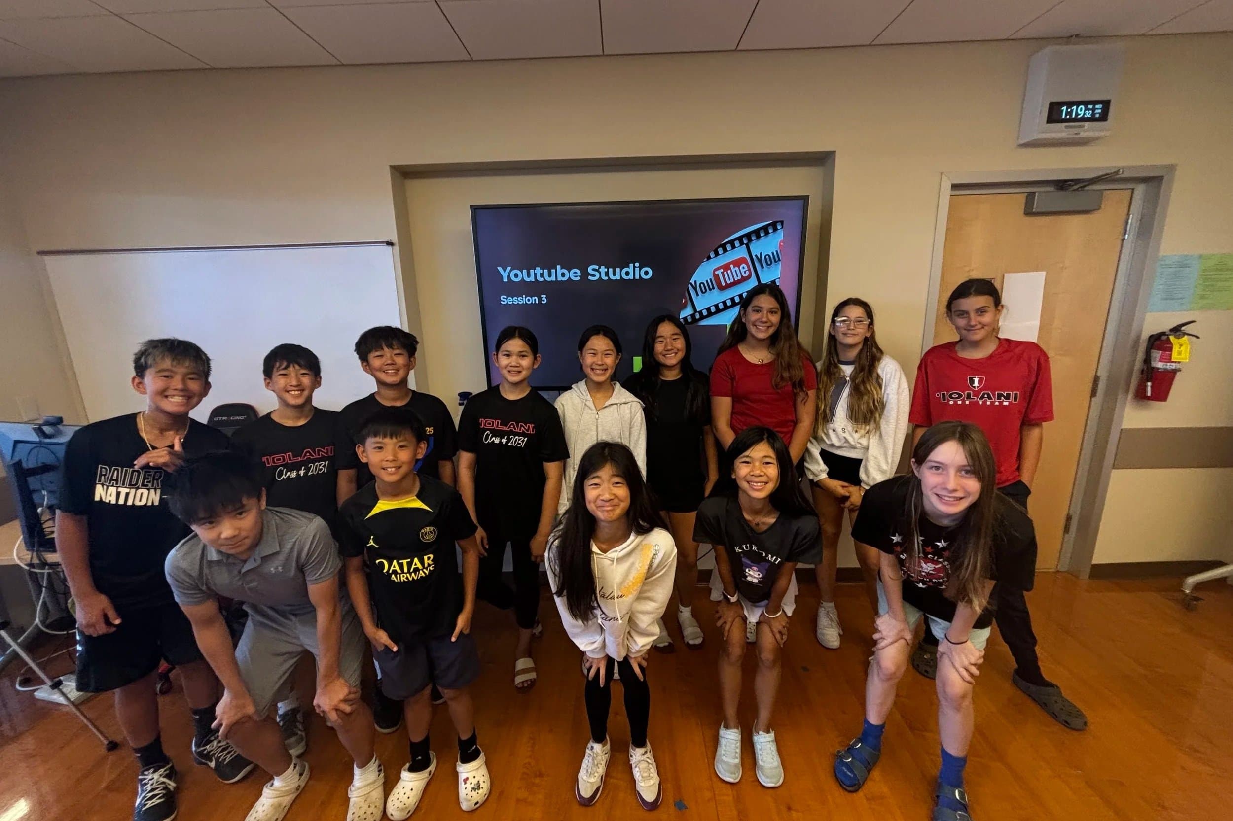 A group of children and teenagers standing and kneeling in a classroom for a group photo, with a digital screen behind them displaying 'Youtube Studio Session 3'.