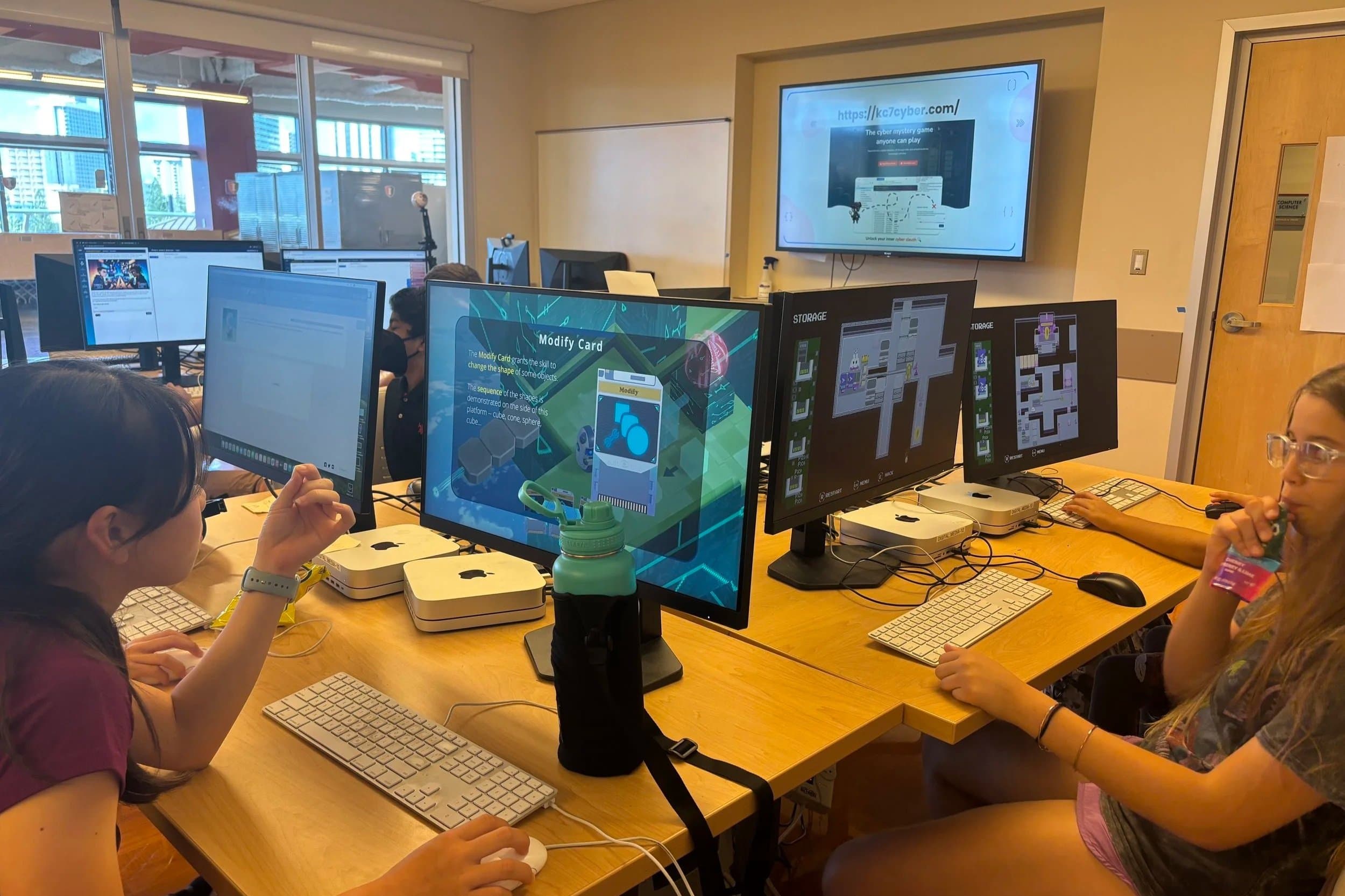 Students working at computers in a classroom with a wall-mounted screen displaying a website.