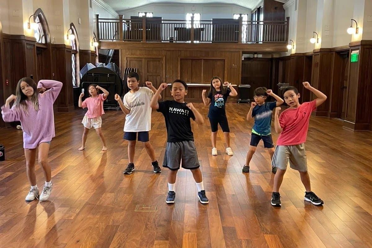 Seven children in casual clothes dancing or exercising on a wooden floor in a large room with wood-paneled walls and a balcony.