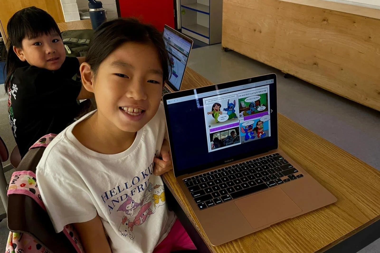 Smiling young girl showing her laptop with a comic strip on the screen, sitting at a wooden desk in a classroom, with another girl in the background.