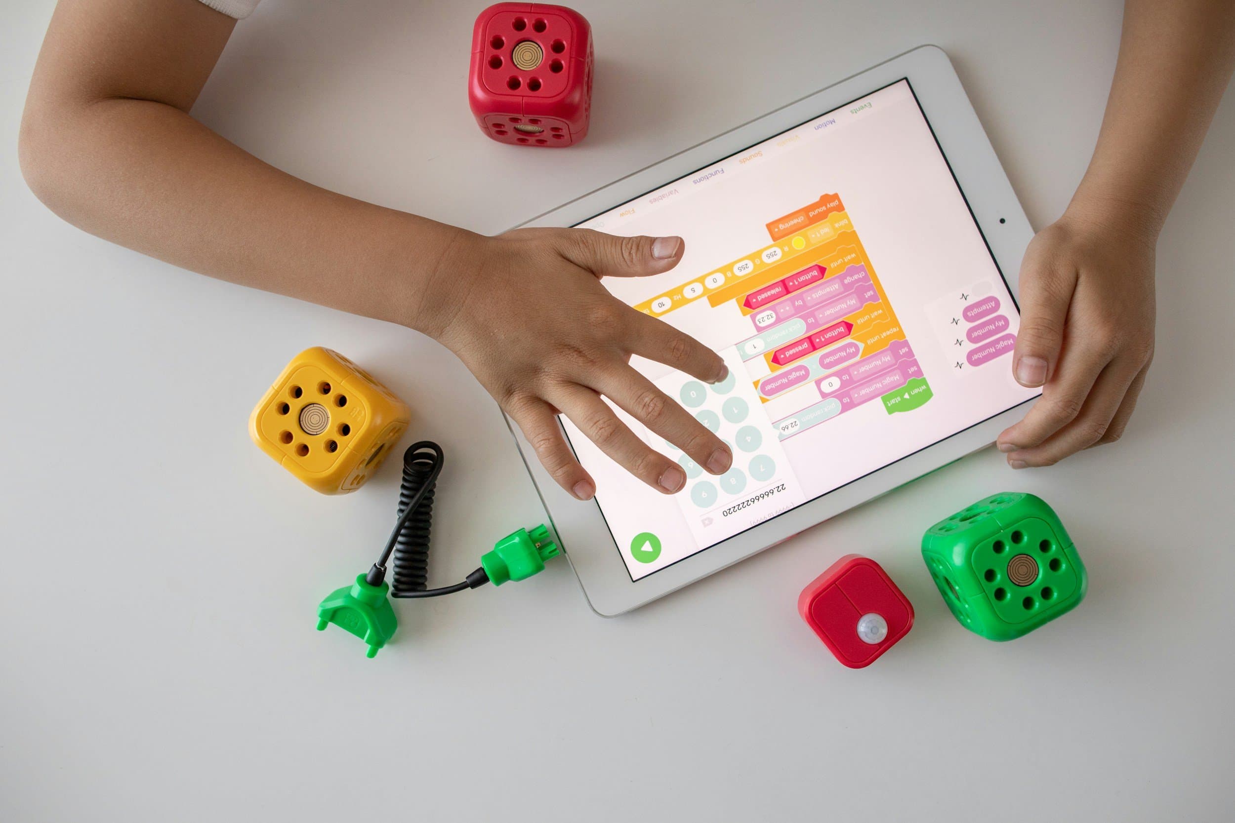 A child interacts with a tablet displaying a block-based coding interface. The tablet is surrounded by colorful toy dice and a coiled electronic wire with a connector.