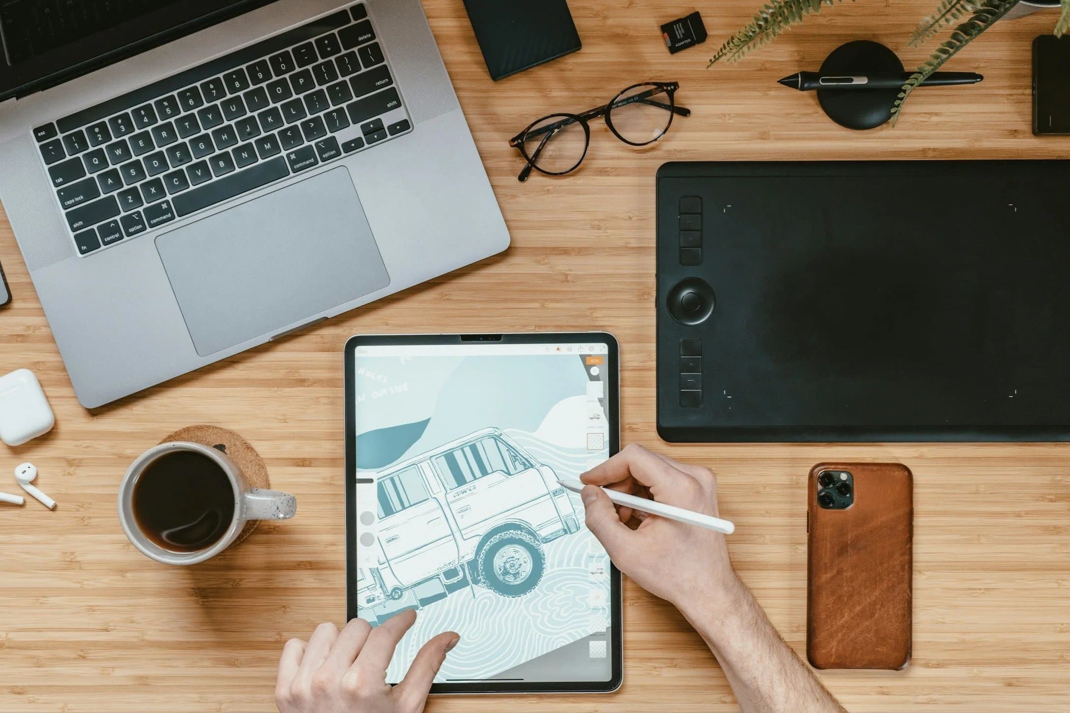 Overhead view of a workspace with a silver laptop, a black graphics tablet, a tablet displaying a digital drawing of an off-road vehicle, a cup of coffee, a pair of glasses, wireless earbuds, a brown smartphone, and various small office supplies on a wooden surface.