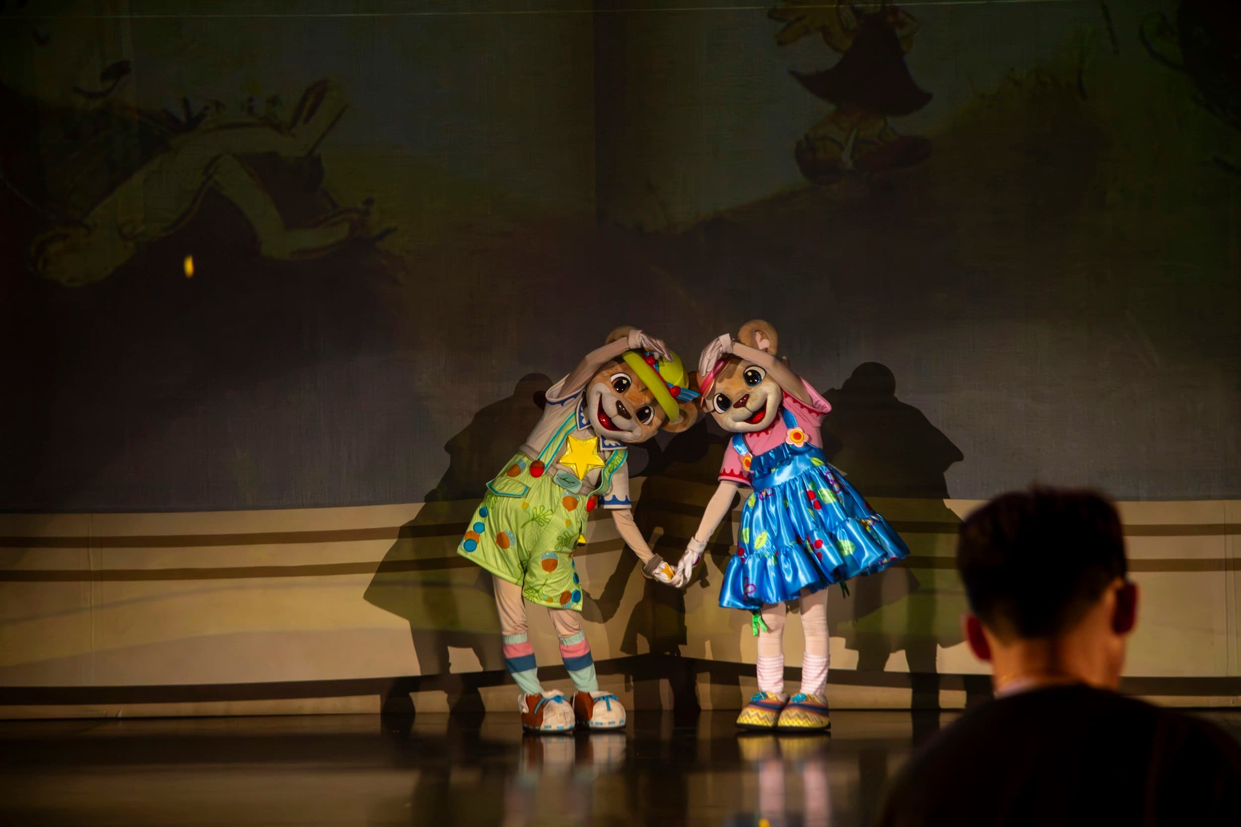 Two costumed characters in bright colorful costumes, holding hands and standing on a stage with a dark backdrop and faint images of animals in the background.