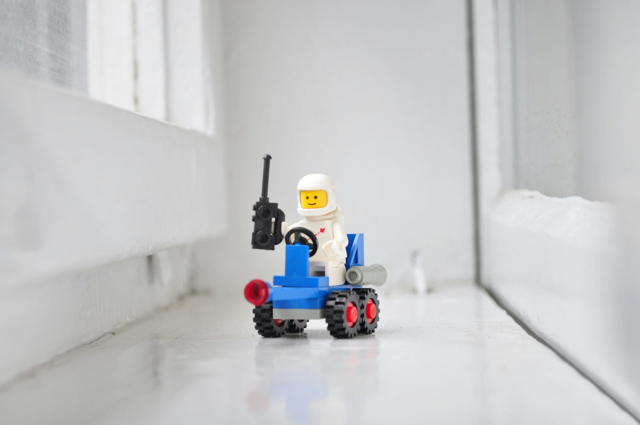 LEGO astronaut figurine driving a small blue and gray construction vehicle with a black radio or walkie-talkie, positioned on a white surface near a white wall and window.